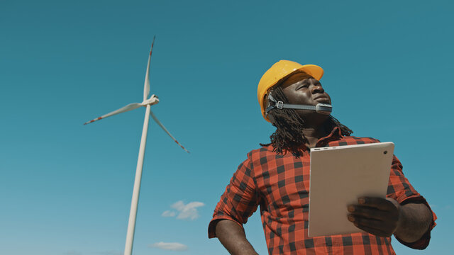 Adult or mature professional african man in hard hat standing with modern digital tablet against win