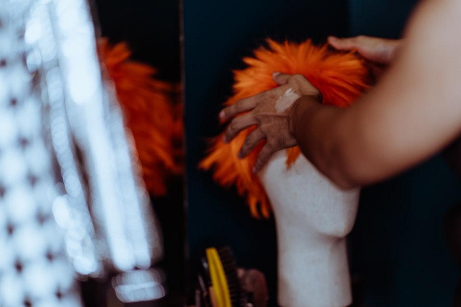 Crop anonymous ethnic person preparing fake wig on mannequin while dressing on room near mirror