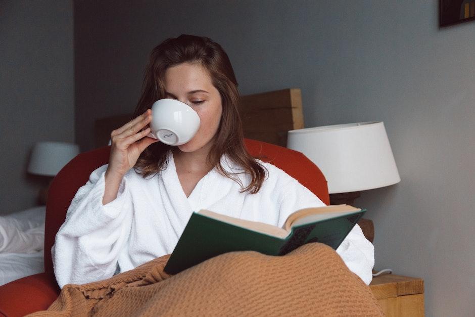 Woman in White Robe Drinking on White Ceramic Mug while Reading a Book